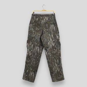May include: Camouflage cargo pants with a green, brown, and white pattern. The pants have multiple pockets and are hanging on a gold hanger. The fabric features a leaf and tree bark design.
