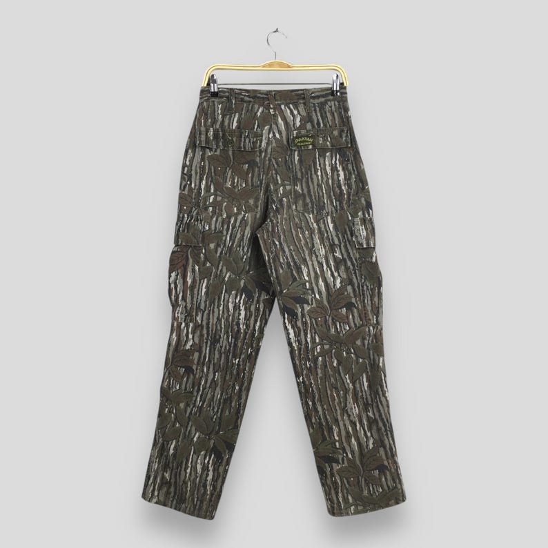 May include: Camouflage cargo pants with a green, brown, and white pattern. The pants have multiple pockets and are hanging on a gold hanger. The fabric features a leaf and tree bark design.