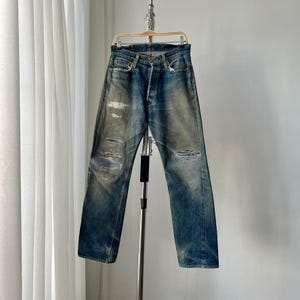 Puede incluir: Vaqueros de mezclilla azul descolorido con detalles desgastados, incluyendo rasgaduras y parches en las rodillas. Los vaqueros cuelgan de una percha sobre un fondo neutro, mostrando una estética vintage o desgastada.