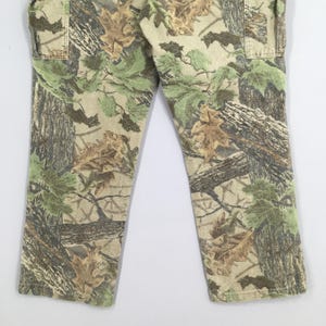 May include: Camouflage pants featuring a realistic forest pattern with shades of green, brown, and tan. The pants have a classic cut with pockets and a neutral background. The fabric appears to be durable, suitable for outdoor activities.