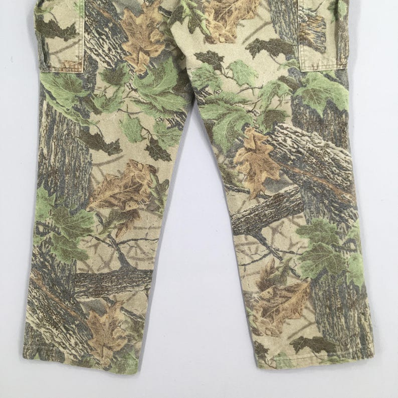 May include: Camouflage pants featuring a realistic forest pattern with shades of green, brown, and tan. The pants have a classic cut with pockets and a neutral background. The fabric appears to be durable, suitable for outdoor activities.