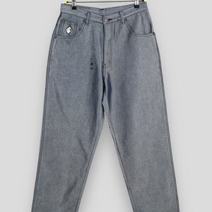 Könnte beinhalten: Eine hellgraue Jeanshose mit einem kleinen Logo-Patch auf der rechten Tasche. Die Jeans hängt an einem Holzbügel.
