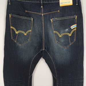 May include: Dark wash denim jeans with yellow stitching and pocket detailing. The back pockets feature a unique design with yellow stitching. A tag with the text "XV-FUNCTION" is attached to the waistband.