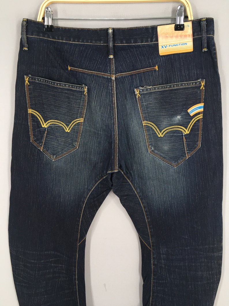 May include: Dark wash denim jeans with yellow stitching and pocket detailing. The back pockets feature a unique design with yellow stitching. A tag with the text "XV-FUNCTION" is attached to the waistband.