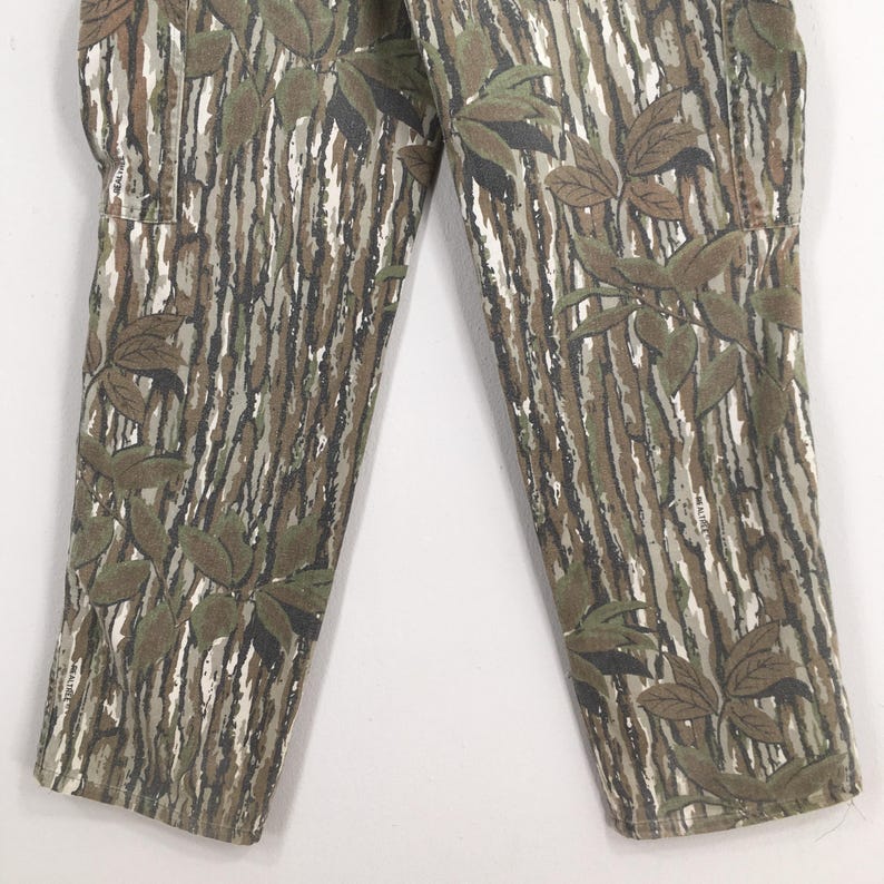 May include: Close-up of camouflage pants featuring a realistic tree bark pattern in shades of brown, green, and white. The pants have a leaf design overlaying the bark pattern. The fabric appears to be a durable material.