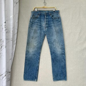 Könnte beinhalten: Verblasste blaue Jeans aus Denim, die an einem goldfarbenen Kleiderbügel hängen. Die Jeans haben einen Knopfverschluss und ein klassisches Fünf-Taschen-Design. Der Denim weist Gebrauchsspuren und Ausbleichungen auf, was ihm einen Vintage-Look verleiht. Die Jeans sind im Straight-Leg-Stil.