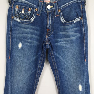 May include: Blue denim jeans with distressed details, featuring orange stitching and decorative pocket stitching. The jeans have a button closure and a classic five-pocket design. The True Religion logo is visible on the back pocket.