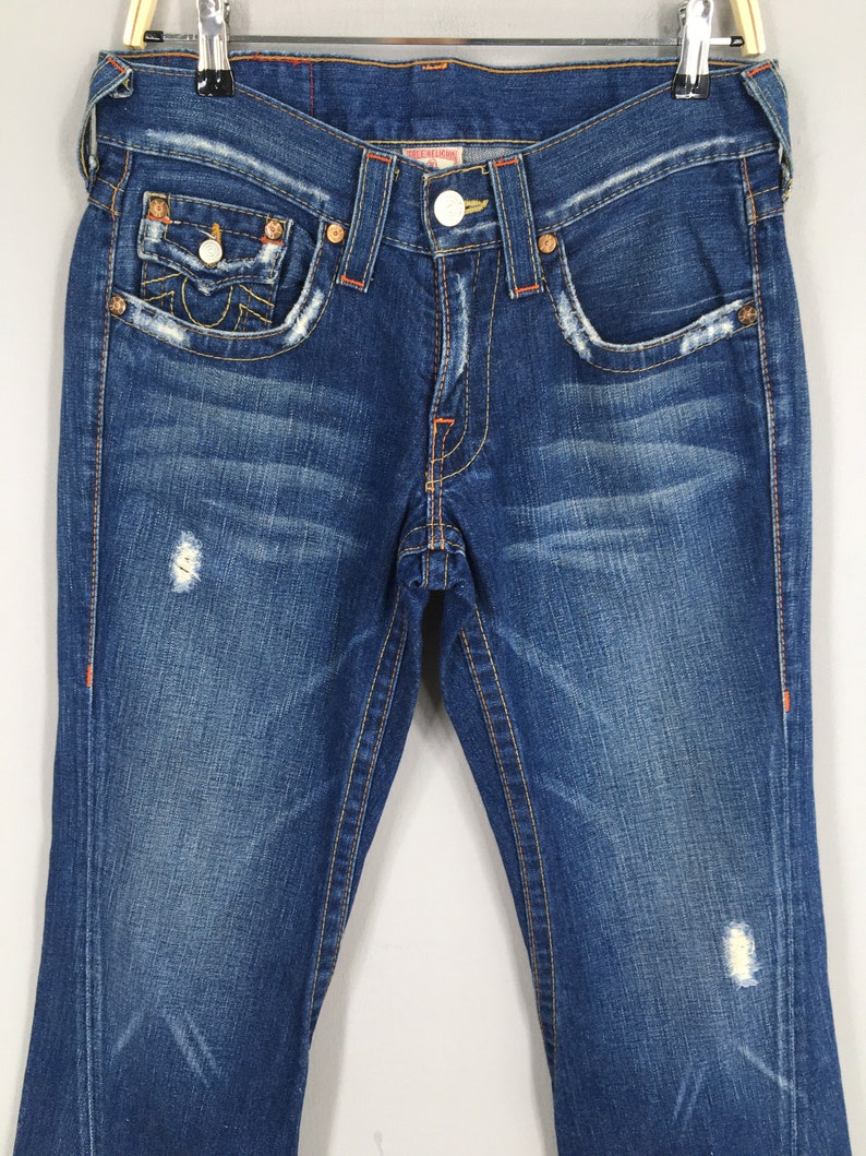 May include: Blue denim jeans with distressed details, featuring orange stitching and decorative pocket stitching. The jeans have a button closure and a classic five-pocket design. The True Religion logo is visible on the back pocket.