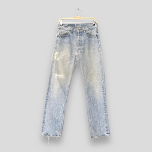 Puede incluir: Vaqueros de mezclilla azul claro con un lavado descolorido y detalles desgastados, colgados de una percha dorada. Los vaqueros tienen bragueta con botones y un diseño clásico de cinco bolsillos. Los vaqueros tienen un área desgastada en la rodilla izquierda.