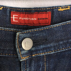 Puede incluir: Primer plano de unos vaqueros de mezclilla azul oscuro. Los vaqueros presentan una etiqueta roja con el texto "FUNCTION EDWIN" y un bot&oacute;n plateado con la letra "E". Se ven costuras amarillas a lo largo de las costuras.