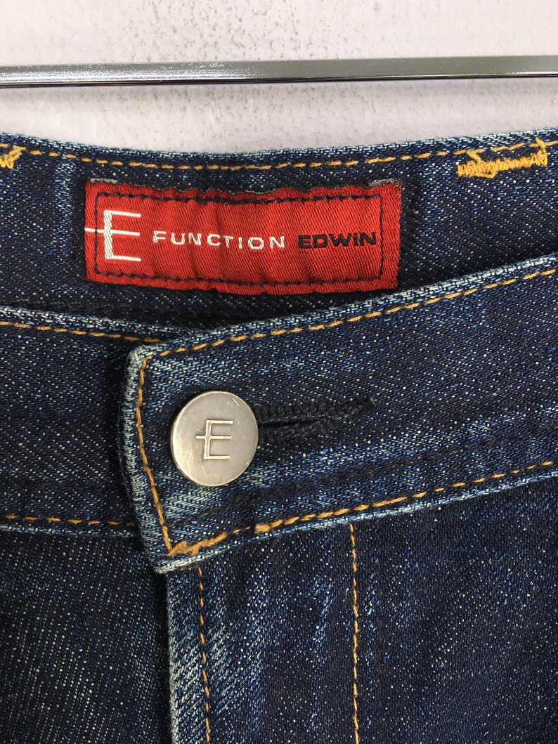 Puede incluir: Primer plano de unos vaqueros de mezclilla azul oscuro. Los vaqueros presentan una etiqueta roja con el texto "FUNCTION EDWIN" y un bot&oacute;n plateado con la letra "E". Se ven costuras amarillas a lo largo de las costuras.