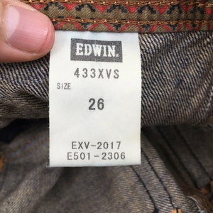 May include: Close-up of a denim tag with the brand name "EDWIN" in white. The tag displays the size "26" and product codes "433XVS", "EXV-2017", and "E501-2306". The tag is set against a denim background with a patterned fabric.