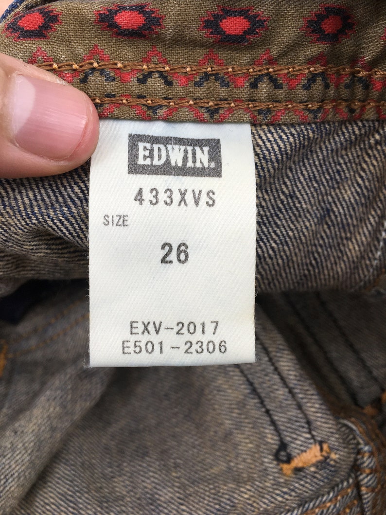 May include: Close-up of a denim tag with the brand name "EDWIN" in white. The tag displays the size "26" and product codes "433XVS", "EXV-2017", and "E501-2306". The tag is set against a denim background with a patterned fabric.