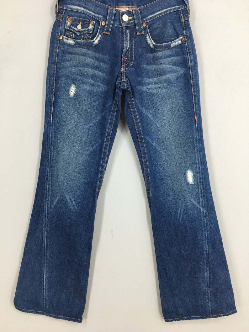 May include: Blue denim jeans with a bootcut leg. The jeans have a distressed look with faded areas and small rips. Orange stitching accents the pockets and seams. The back pockets have a decorative design.