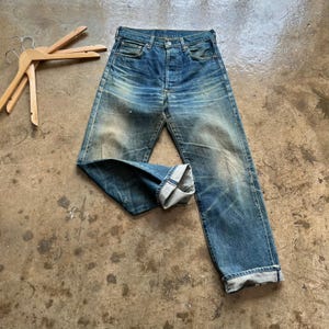Könnte beinhalten: Eine blaue Jeans aus Denim mit verwaschener Optik, Knopfleiste und fünf Taschen. Die Hose hat einen geraden Schnitt mit umgeschlagenen Beinabschlüssen, die den helleren blauen Innenstoff sowie deutliche Abnutzungsspuren und Falten zeigen.