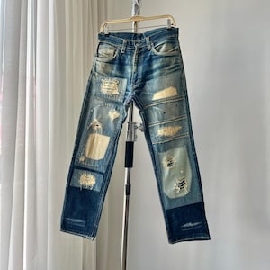 Puede incluir: Vaqueros azules de mezclilla con parches desgastados y detalles rasgados. Los vaqueros tienen un lavado descolorido y cuelgan de una percha de metal. Los parches son en varios tonos de beige y azul.