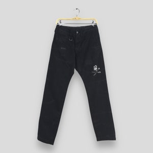 May include: Black pants with a straight leg design, hanging on a gold hanger. The pants feature a small white skull and crossbones graphic on the right leg. A small logo is visible on the left side.