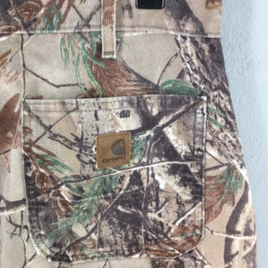 K&ouml;nnte beinhalten: Nahaufnahme einer Carhartt Camouflage-Hose. Die Hose hat ein Wald-Camouflage-Muster in Braun-, Gr&uuml;n- und Beiget&ouml;nen. Eine quadratische Tasche ist sichtbar, zusammen mit einem Carhartt-Markenaufn&auml;her. Die Hose ist aus strapazierf&auml;higem Stoff gefertigt.