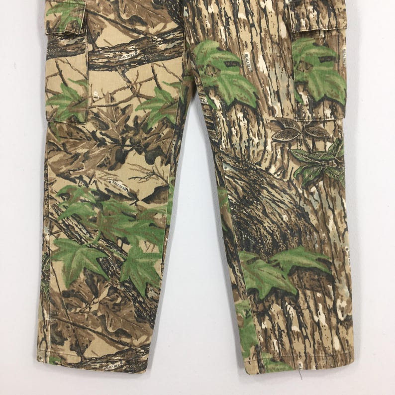 Puede incluir: Primer plano de pantalones cargo de camuflaje. Los pantalones presentan un patr&oacute;n forestal realista con tonos de marr&oacute;n, beige y verde. El dise&ntilde;o incluye patrones de hojas y corteza de &aacute;rbol. Los pantalones tienen bolsillos y cierre de bot&oacute;n.
