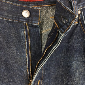 Puede incluir: Primer plano de unos vaqueros de mezclilla azul oscuro con una cremallera parcialmente desabrochada. Los vaqueros tienen un bot&oacute;n plateado y una etiqueta roja que dice "FUNCTION EDWIN". Las costuras son de color amarillo contrastante.
