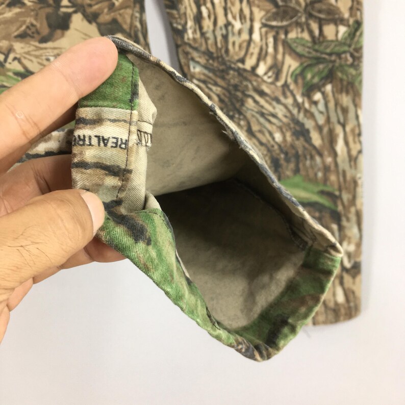 Puede incluir: Primer plano de pantalones de camuflaje con una etiqueta Realtree visible. Los pantalones presentan un patr&oacute;n realista de marr&oacute;n, verde y beige, que imita un entorno forestal. La tela est&aacute; doblada, revelando el forro interior.