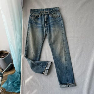 Könnte beinhalten: Eine blaue Jeans aus Denim mit verwaschener Optik, ausgestattet mit einer Knopfleiste, fünf Taschen und einem geradlinigen Schnitt. Die Beinabschlüsse sind umgeschlagen und zeigen ein weiß-blaues Selvedge-Detail an der inneren Naht des Stoffes.