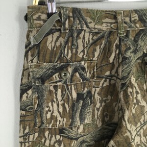 Pu&ograve; includere: Primo piano di pantaloni mimetici con tasca abbottonata. I pantaloni presentano un motivo a corteccia d'albero marrone, verde e beige. Un passante in tessuto verde &egrave; attaccato alla cintura. I pantaloni sono appesi a una gruccia di metallo.