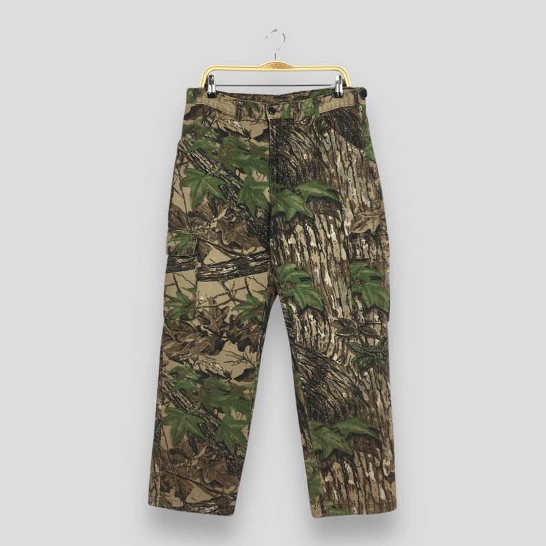Puede incluir: Pantalones cargo de camuflaje con cierre de bot&oacute;n y trabillas para el cintur&oacute;n. Los pantalones presentan un estampado de hojas verdes y corteza de &aacute;rbol marr&oacute;n, con dos bolsillos cargo en los laterales. Los pantalones est&aacute;n colgados de una percha dorada.