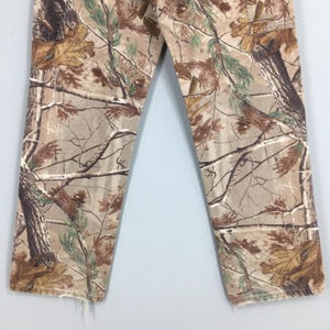 K&ouml;nnte beinhalten: Tarnhose in Braun-, Gr&uuml;n- und Beiget&ouml;nen mit einem realistischen Baum- und Bl&auml;ttermuster. Die Hose hat einen geraden Beinverlauf und einen ausgefransten Saum. Der Stoff scheint ein strapazierf&auml;higes Material zu sein, das f&uuml;r Outdoor-Aktivit&auml;ten geeignet ist.