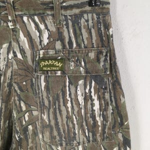 May include: Close-up of camouflage pants with a pocket. The pants feature a pattern of brown, green, and white vertical stripes. A green patch on the pocket reads "Spartan Realtree".