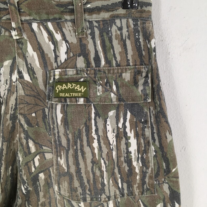 May include: Close-up of camouflage pants with a pocket. The pants feature a pattern of brown, green, and white vertical stripes. A green patch on the pocket reads "Spartan Realtree".