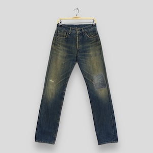 May include: Vintage blue denim jeans with a faded wash and a patched knee. The jeans are hanging on a gold-colored hanger. The denim has a worn appearance with subtle distressing.