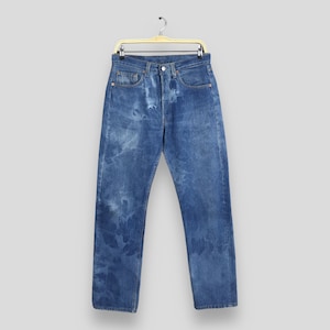 May include: Blue denim jeans with a faded, distressed wash. The jeans are straight-legged and feature classic five-pocket styling. They are hanging on a gold-colored hanger against a neutral background.