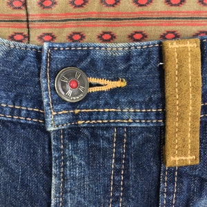 May include: Close-up of blue denim jeans with a decorative button and a brown suede belt loop. The button is silver with a red center. The jeans have yellow stitching and a patterned fabric lining.