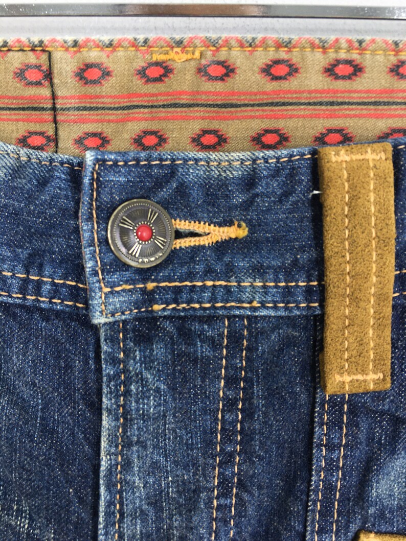 May include: Close-up of blue denim jeans with a decorative button and a brown suede belt loop. The button is silver with a red center. The jeans have yellow stitching and a patterned fabric lining.