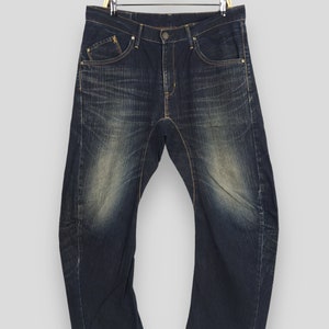 May include: Dark wash denim jeans with a faded, worn appearance. The jeans have a classic five-pocket design, a button and zipper closure, and are hanging on a gold-colored hanger. The denim has a distressed look with lighter areas on the thighs.