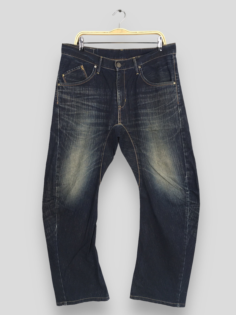 May include: Dark wash denim jeans with a faded, worn appearance. The jeans have a classic five-pocket design, a button and zipper closure, and are hanging on a gold-colored hanger. The denim has a distressed look with lighter areas on the thighs.