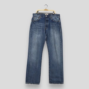 May include: A pair of blue denim jeans hanging on a wooden hanger. The jeans have a button fly and five pockets.