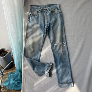 Könnte beinhalten: Hellblaue Jeans aus Denim mit verwaschener Optik, Knopfleiste, fünf Taschen und einem Riss am linken Knie. Die Hose mit geradem Bein hat umgeschlagene Säume und weist leichte Gebrauchsspuren sowie eine dezente Verfärbung auf.