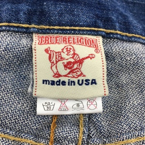 May include: Close-up of a denim garment label. The label is rectangular with the text "TRUE RELIGION" in red. Below the text is a red graphic of a person playing a guitar. The words "made in USA" are printed below the graphic.