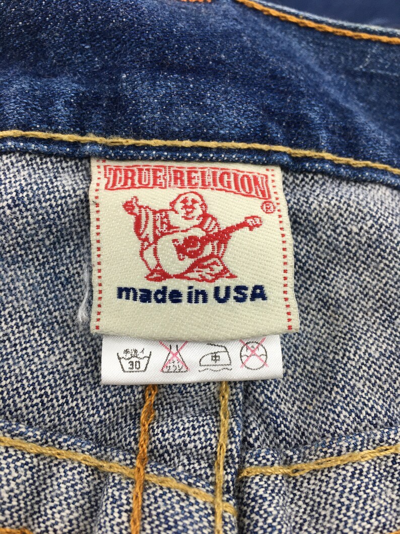 May include: Close-up of a denim garment label. The label is rectangular with the text "TRUE RELIGION" in red. Below the text is a red graphic of a person playing a guitar. The words "made in USA" are printed below the graphic.