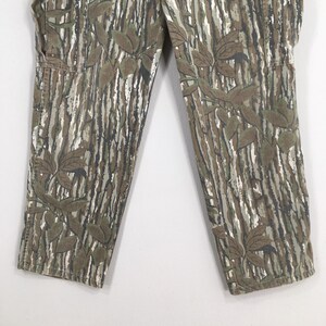 May include: Close-up of camouflage pants with a leaf pattern. The pants feature a vertical striped design in shades of brown, green, and white, with darker leaf accents. The fabric appears to be a durable material, suitable for outdoor wear.