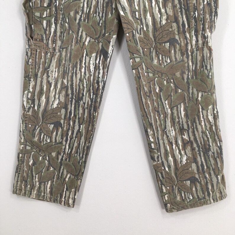 May include: Close-up of camouflage pants with a leaf pattern. The pants feature a vertical striped design in shades of brown, green, and white, with darker leaf accents. The fabric appears to be a durable material, suitable for outdoor wear.