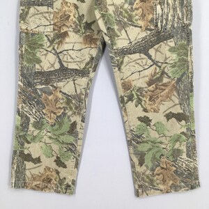 May include: Camouflage-patterned pants in shades of green, brown, and beige. The pants feature a classic camouflage design with a mix of leaves and tree branch patterns. The fabric appears to be a durable material, suitable for outdoor activities.