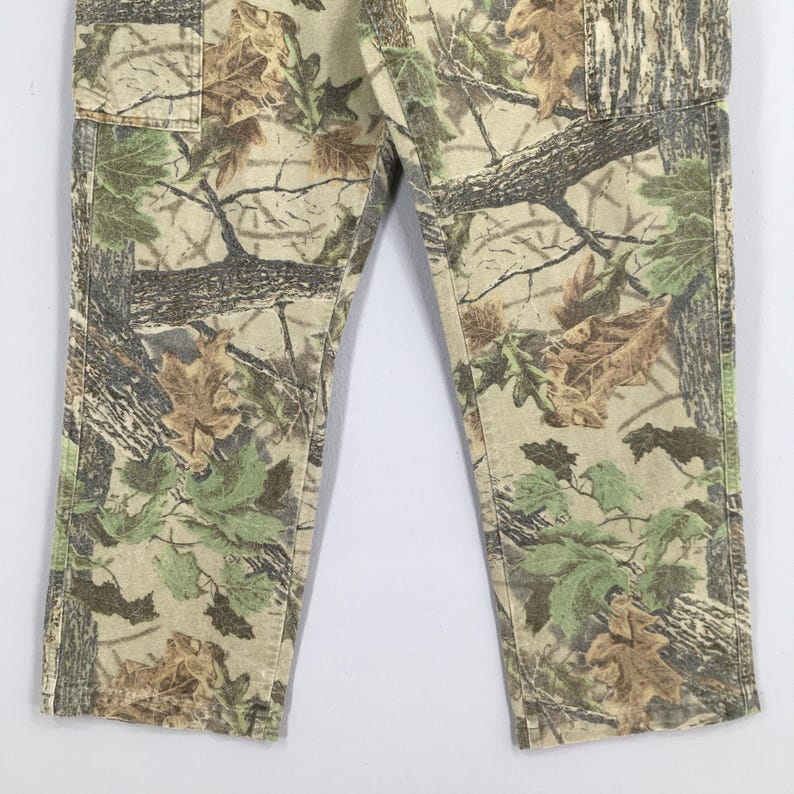 May include: Camouflage-patterned pants in shades of green, brown, and beige. The pants feature a classic camouflage design with a mix of leaves and tree branch patterns. The fabric appears to be a durable material, suitable for outdoor activities.