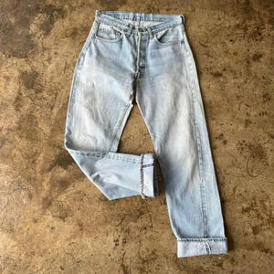 Puede incluir: Vaqueros de mezclilla azul claro con bragueta de botones y puños doblados. Los vaqueros tienen un diseño clásico de cinco bolsillos y un lavado descolorido, lo que les da un aspecto vintage. Los vaqueros están colocados sobre una superficie texturizada.