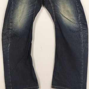 May include: Close-up of a pair of dark blue denim jeans. The jeans have a faded, worn look with lighter patches on the thighs and knees. Yellow stitching is visible along the seams and hems.