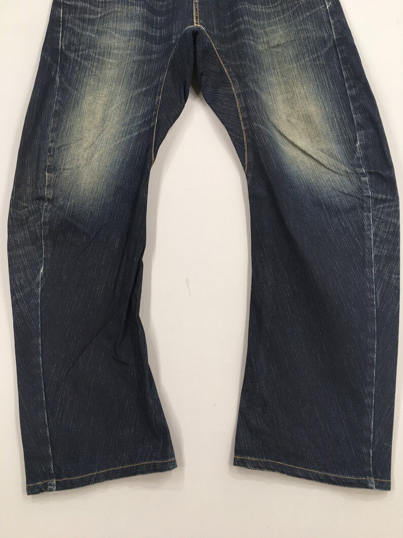 May include: Close-up of a pair of dark blue denim jeans. The jeans have a faded, worn look with lighter patches on the thighs and knees. Yellow stitching is visible along the seams and hems.