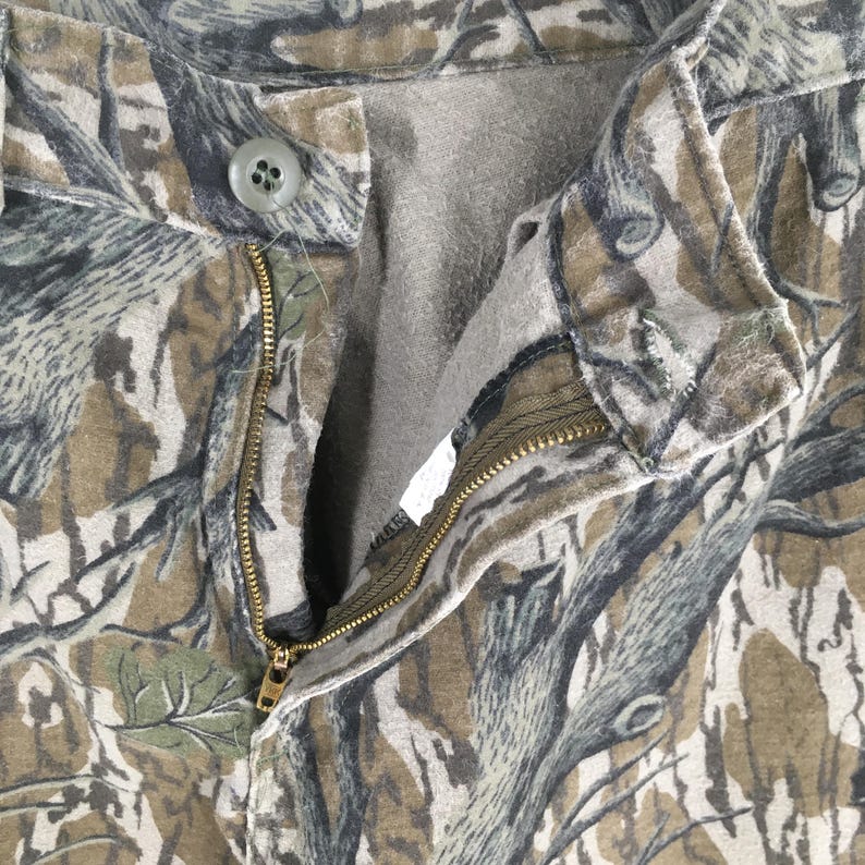 Pu&ograve; includere: Primo piano di pantaloni mimetici con chiusura a bottone e cerniera. I pantaloni presentano un motivo marrone, verde e grigio, che ricorda una scena di foresta. La cerniera &egrave; dorata.