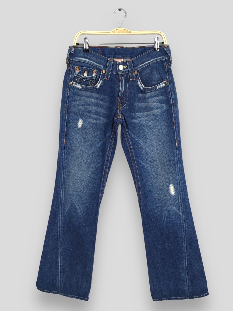 May include: Dark blue denim jeans with a distressed look, featuring a classic five-pocket design. The jeans have a slight flare at the bottom and are hanging on a gold-colored hanger. Orange stitching accents the seams and pockets.